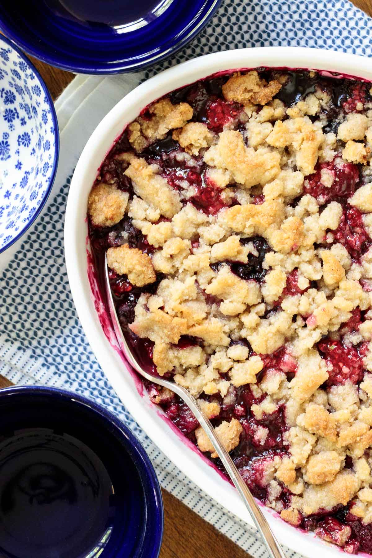 Overhead vertical closeup photo of a Ridiculously Easy Summer Berry Crisp in a white porcelain serving dish surrounded by blue and white individual serving dishes.