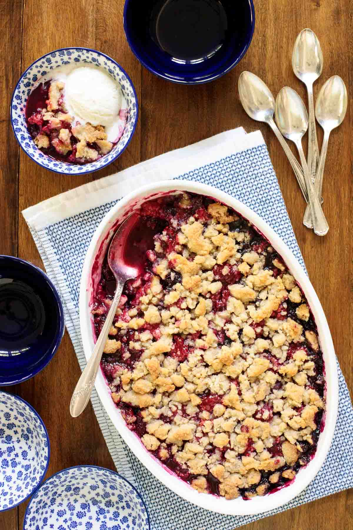 Overhead vertical photo of a Ridiculously Easy Summer Berry Crisp surrounded by serving utensils and individual bowls with ice cream on a wooden table.