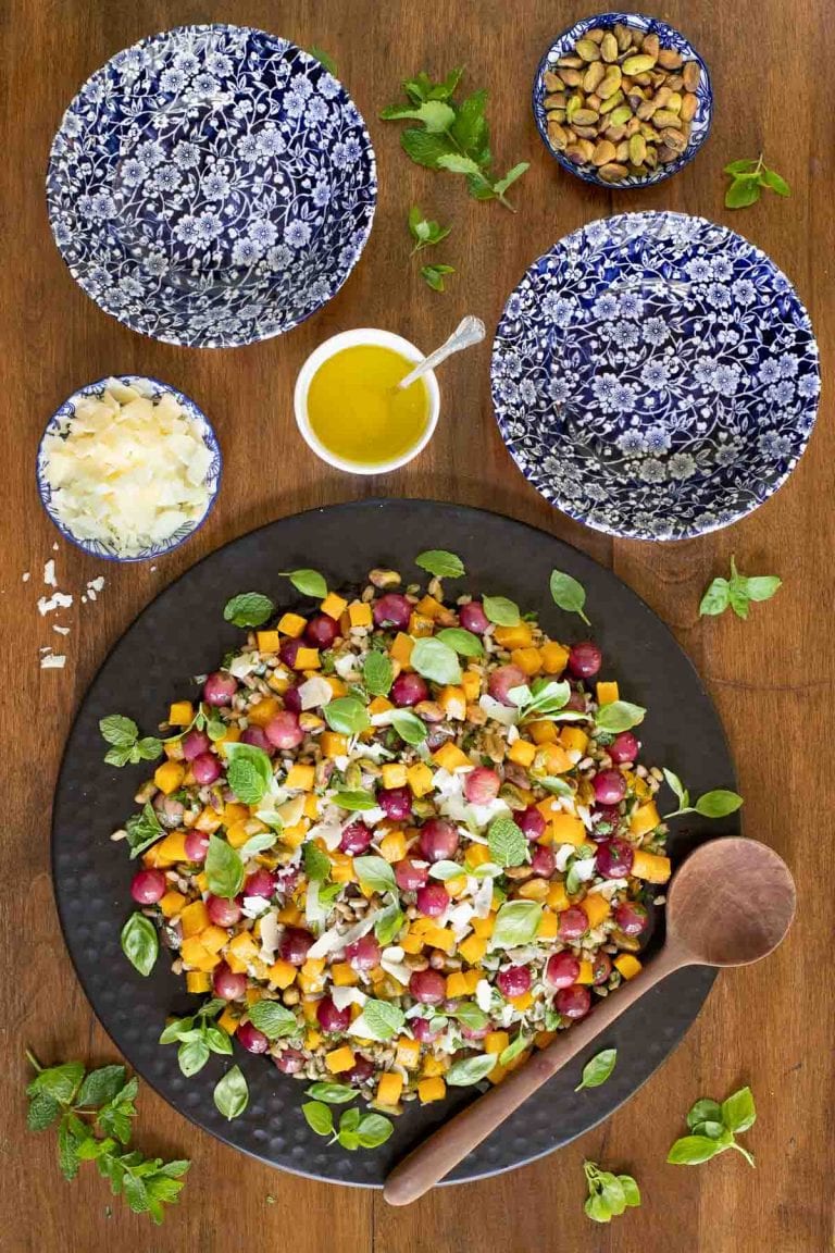 Vertical picture of Roasted Butternut Farro Salad in a black bowl on a wooden table with cheese, nuts and dressing in small bowls surrounding it.
