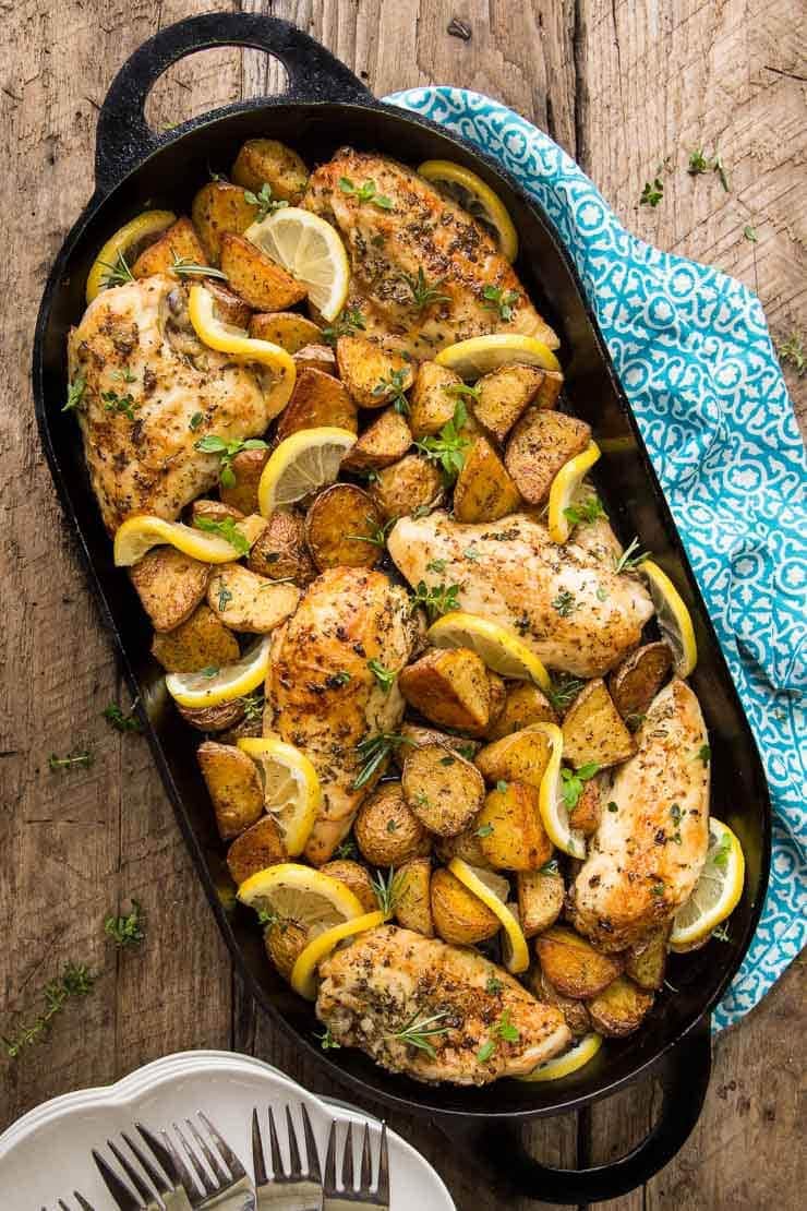 Overhead photo of a cast iron oval baking pan full of Lemon Garlic Roasted Chicken and Potatoes on an aged wood table with a turquoise and white napkin under the pan.