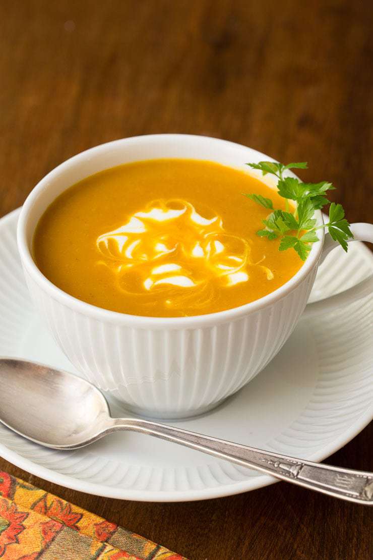 Photo of a white cup filled with Easy Roasted Pumpkin Coconut Curry Soup on a wood table with a spoon and napkin nearby.