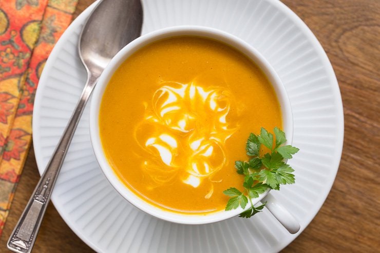 Overhead photo of a cup of Roasted Pumpkin Coconut Curry Soup on a white plate with a napkin and spoon next to it. Garnished with cilantro and a swirl of greek yogurt.