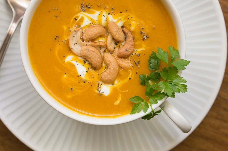 Overhead photo of a cup of Roasted Pumpkin Coconut Curry Soup on a white plate garnished with cilantro, a swirl of greek yogurt and cashew halves.