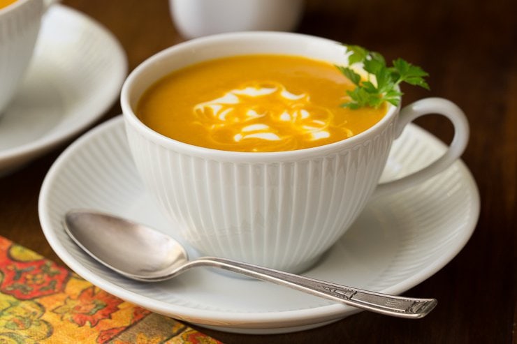 Photo of a white cup filled with Roasted Pumpkin Coconut Curry Soup on a wood table with a fall colored napkin in the foreground.