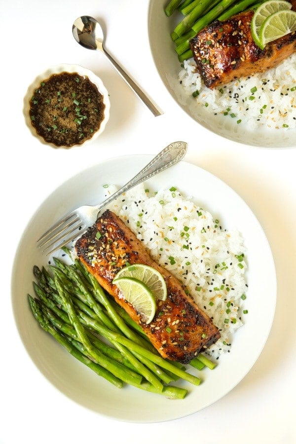 Overhead photo of Honey Ginger Glazed Salmon in white bowls on a bed of seasoned white rice and a side of asparagus. A dish of drizzling sauce and spoon is between the dishes.