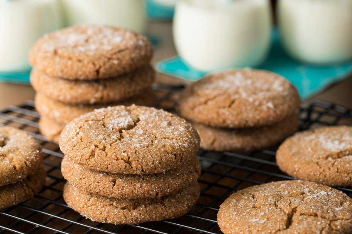 Photo of stacks of Sea Salted Brown Sugar Cookies on a black cooling rack with glasses of milk on turquoise napkins in the background.