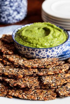 Vertical photo of a batch of Pine Nut Seeded Crackers on an appetizer plate with a dish of avocado dip.