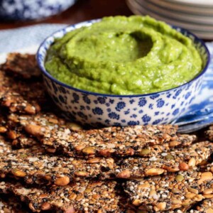 Vertical photo of a batch of Pine Nut Seeded Crackers on an appetizer plate with a dish of avocado dip.