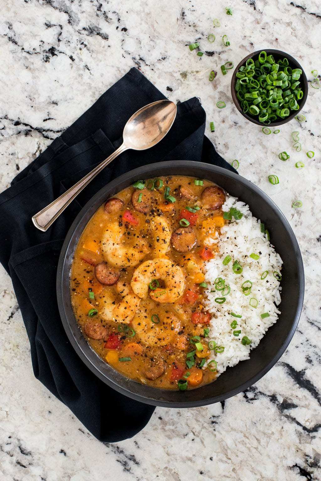 Overhead photo of a bowl of Shrimp and Andouille Gumbo on a granite countertop with a small bowl of green onions and a black napkin.