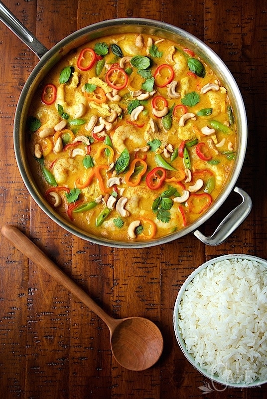 An overhead photo of a sauce pan of Shrimp and Chicken Cashew Curry next to a wood stirring spoon and a bowl of white rice.