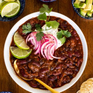 Overhead vertical photo of Slow Roasted Short Rib Chili in a white serving bowl surrounded by garnishes.