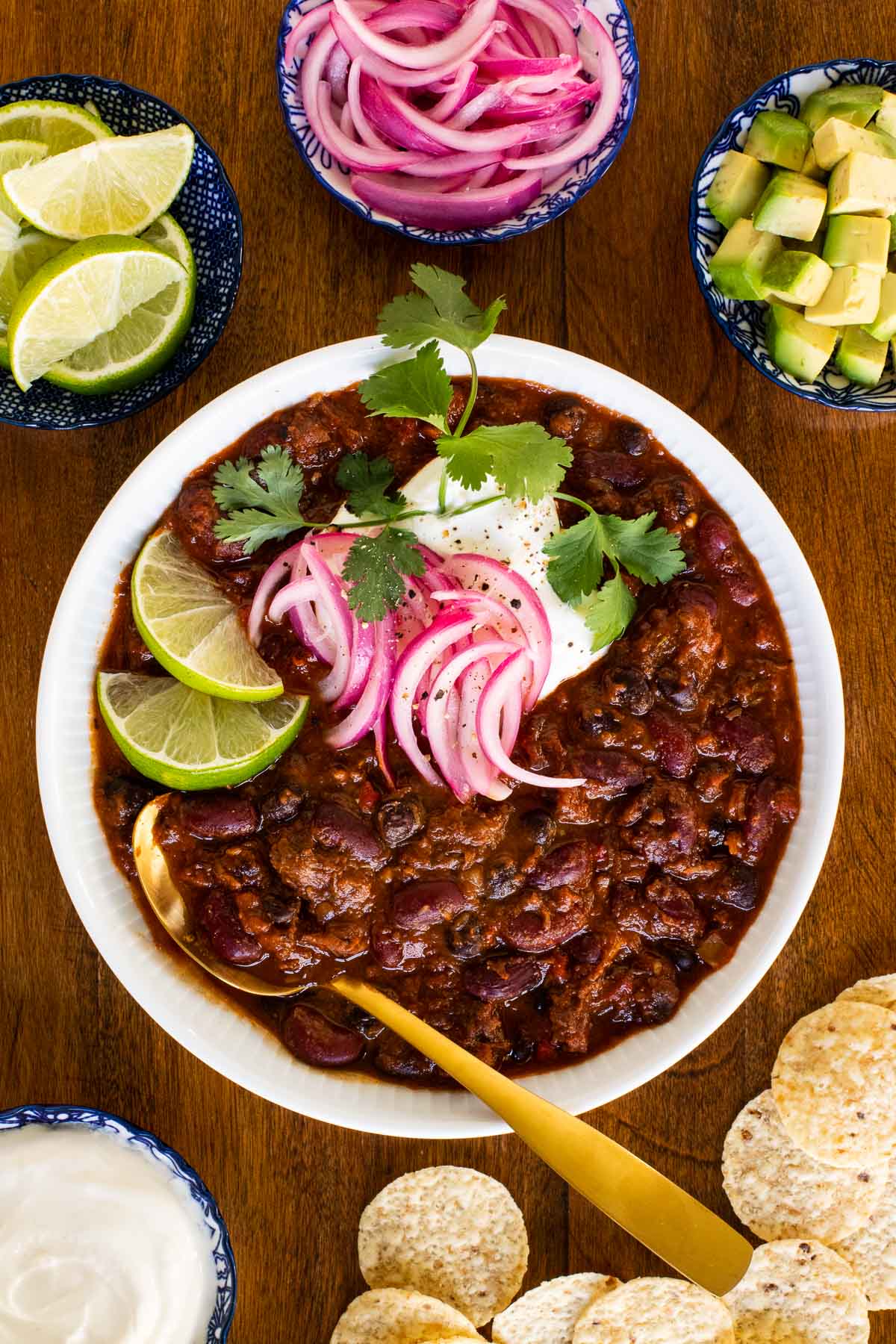 Overhead vertical photo of Slow Roasted Short Rib Chili in a white serving bowl surrounded by sour cream, lime wedges, pickled red onions, diced avocado and chips.
