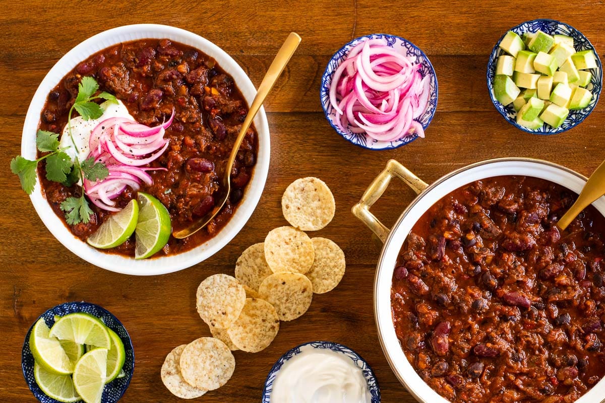 Horizontal overhead photo of a pot and a serving bowl of Slow Roasted Short Rib Chili surrounded by fresh garnishes and tortilla chips.