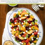 Overhead vertical photo of a serving platter of Stone Fruit Tomato Caprese Salad on a wood table with a small dish of pine nuts and basil vinaigrette dressing.