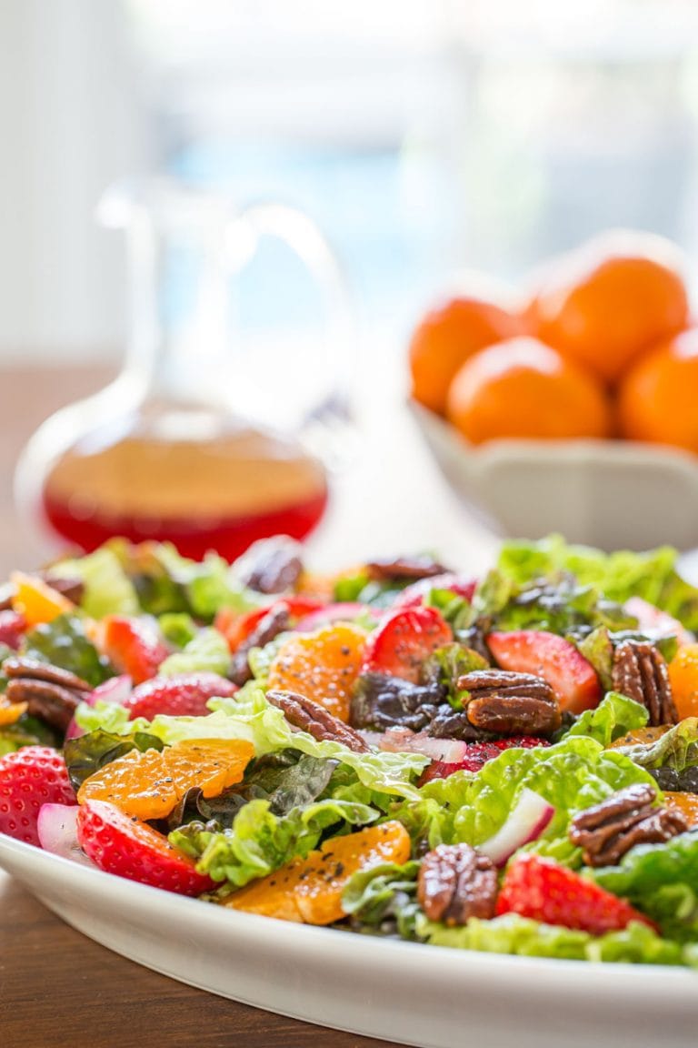 Vertical closeup photo of a Strawberry Clementine Salad on a white platter with dressing in the background.