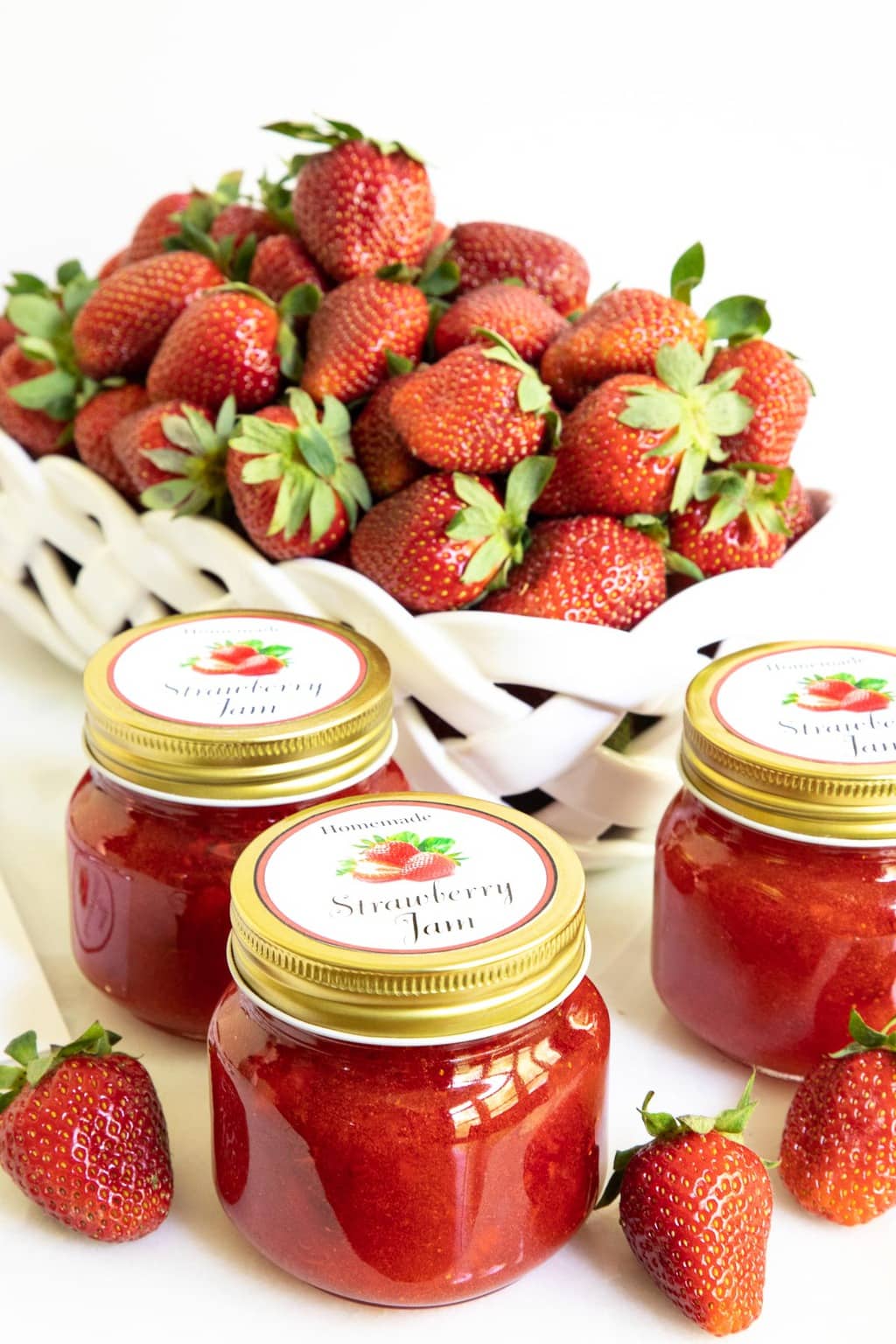Vertical photo of jars of Strawberry Freezer Jam with custom label for gift giving with a white porcelain basket of fresh strawberries in the background.