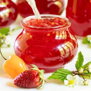 Horizontal closeup photo of a jar of Strawberry Habanero Pepper Jelly with additional jars in the background and a strawberry and a habanero pepper in the foreground.