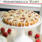 Vertical photo of a Strawberry Jam Shortbread Tart on a white pedestal cake stand on a dining table.
