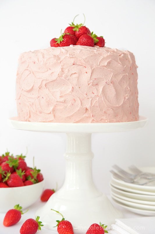 Vertical photo of a Strawberry Layer Cake on a white pedestal cake stand with fresh strawberries and serving utensils and plates in the background.