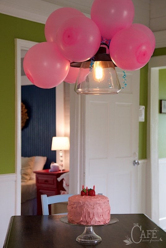 Horizontal photo of a pink birthday cake on a dinning room table with pink balloons attached to a ceiling light fixture.