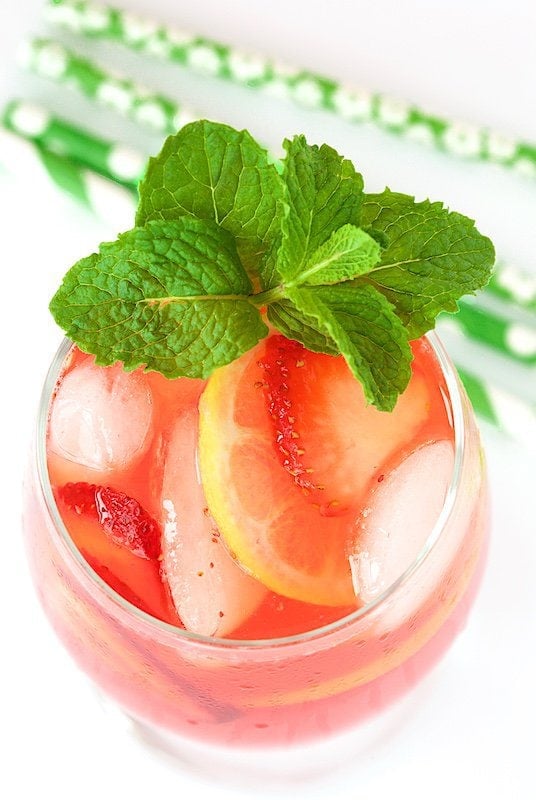 Overhead photo of a glass of Strawberry Lemonade garnished with a sprig of mint and slices of strawberry and lemon. Green straws are in the background.