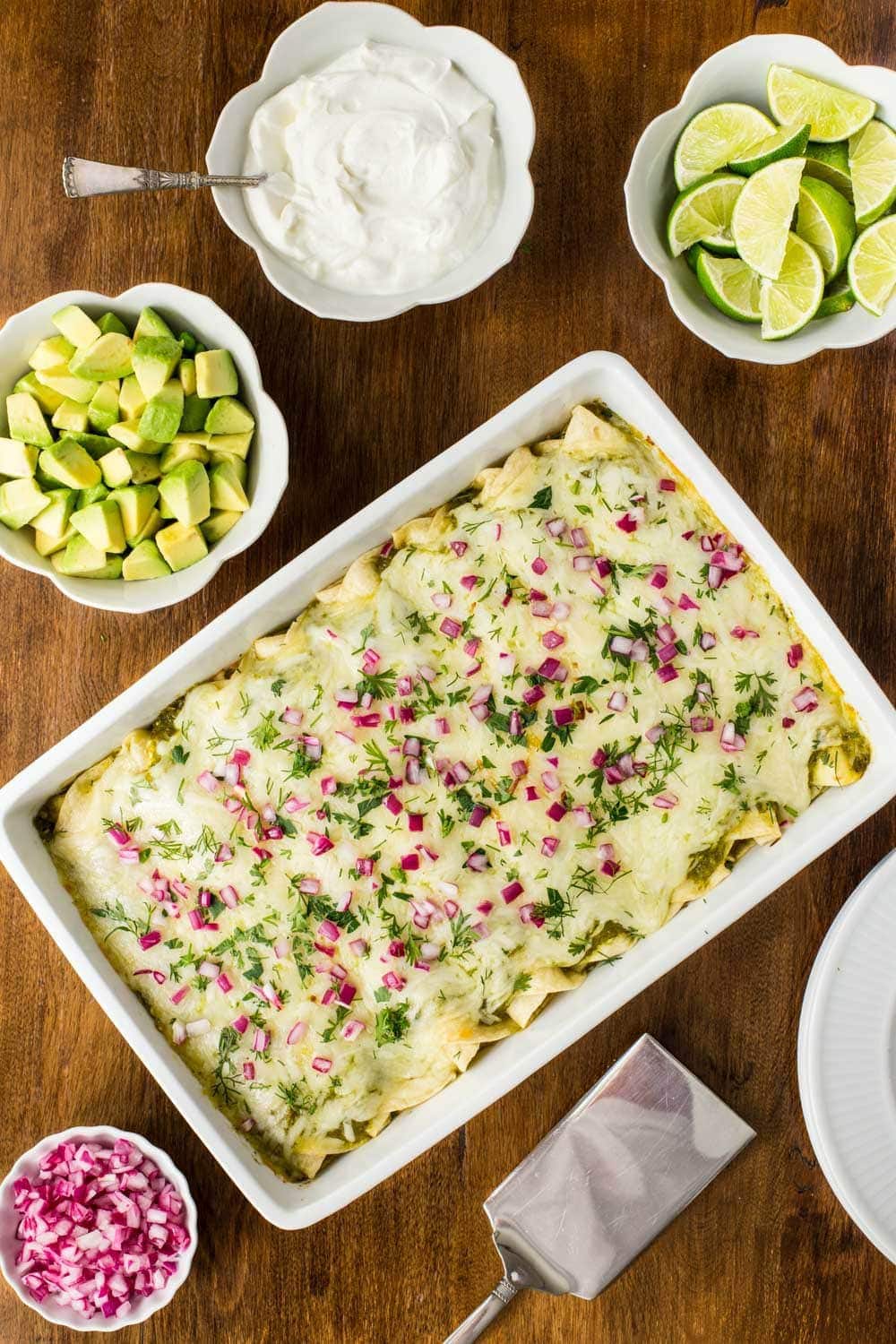 Overhead photo of Super Easy Chicken Enchiladas Verde with all the toppings in separate white bowls surrounding the main dish.