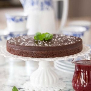Vertical picture of Swedish Chocolate Sticky Cake on a white cake stand with a small glass cruet of raspberry sauce