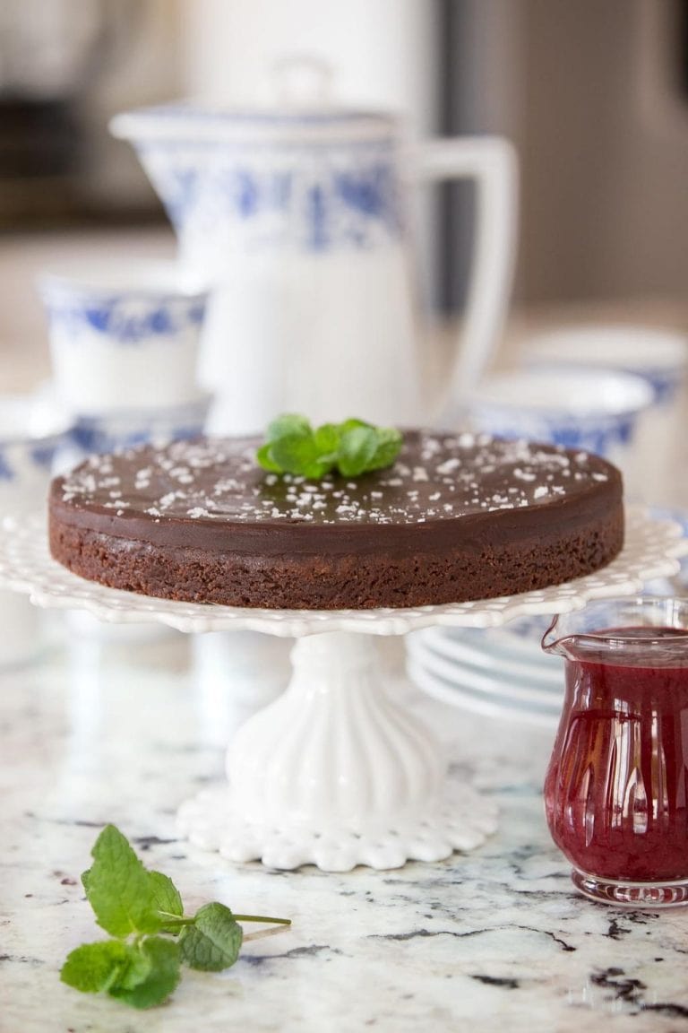 Vertical picture of Swedish Chocolate Sticky Cake on a white cake stand with a small glass cruet of raspberry sauce