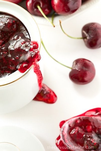 Photo of Sweet Cherry Freezer Jam in a bowl and a spoon with jam drizzling down the side of a white jar filled with jam. 