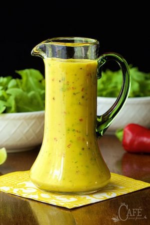 Photo of a glass cruet of Sweet and Spicy Mango Salad Dressing on a yellow and white napkin with bowls of salad and a pepper in the background.