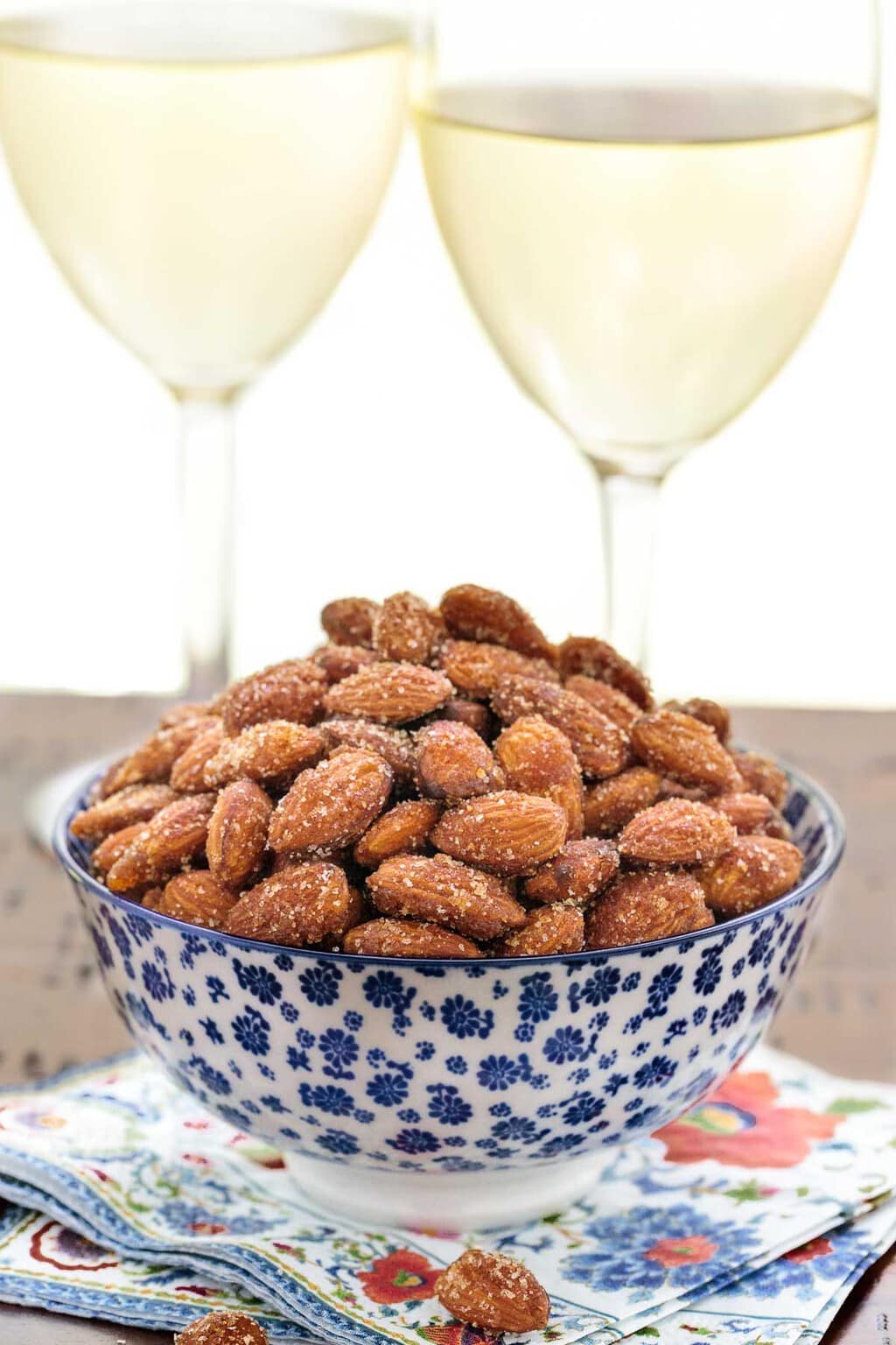 Vertical photo of Sweet and Spicy Almonds in a blue and white patterned bowl in front of two glasses of white wine.