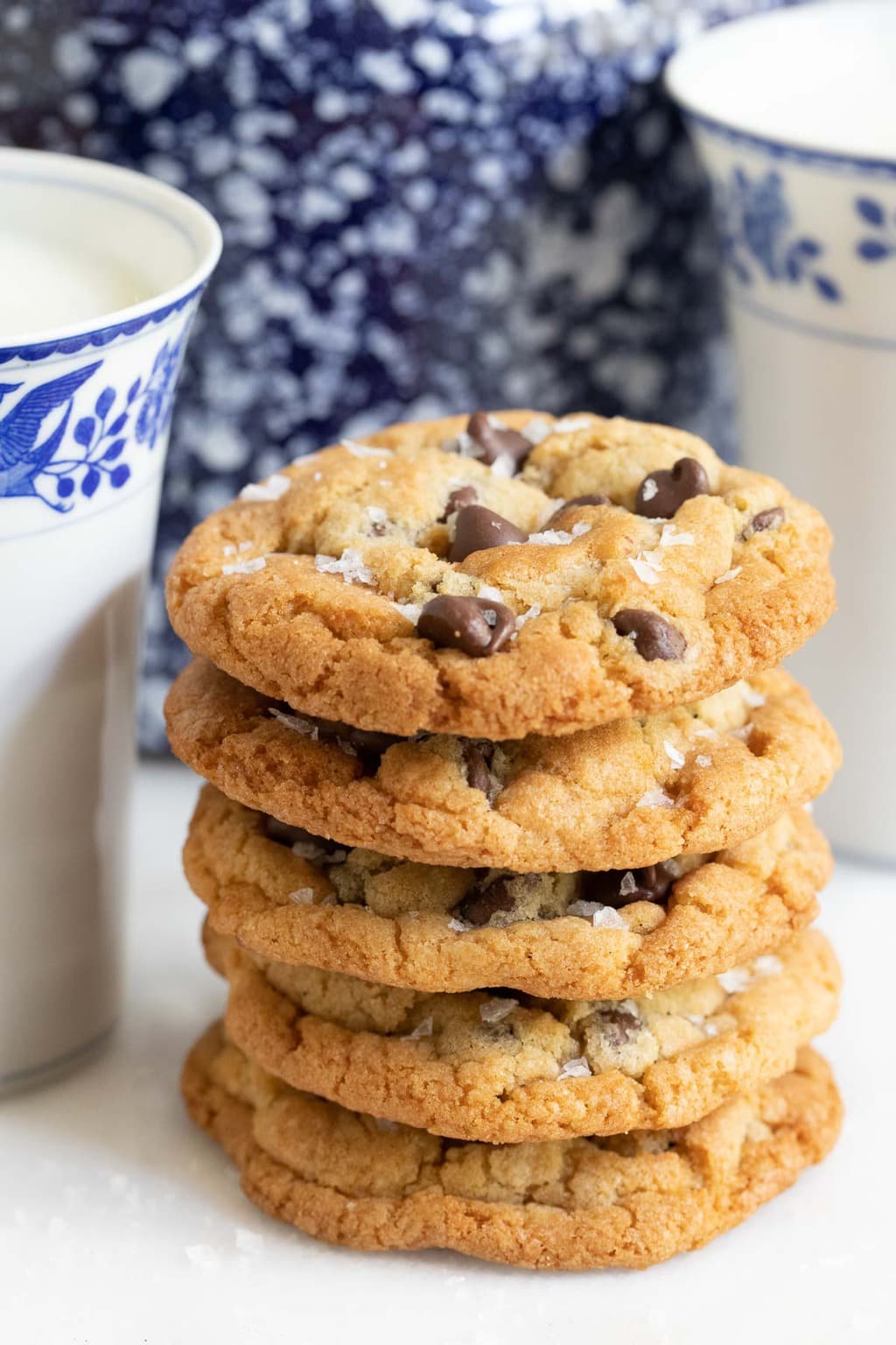 Vertical closeup photo of a stack of The BEST Chocolate Chip Cookies next to two blue and white patterned cups of milk and a cookie jar.