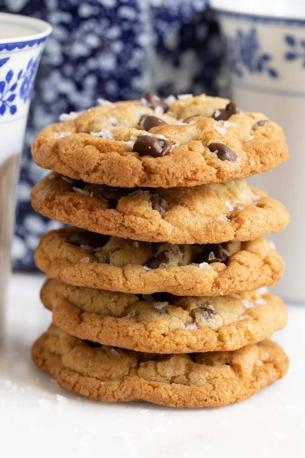 Vertical closeup photo of a stack of The BEST Chocolate Chip Cookies with a cookie jar and milk glasses in the background.
