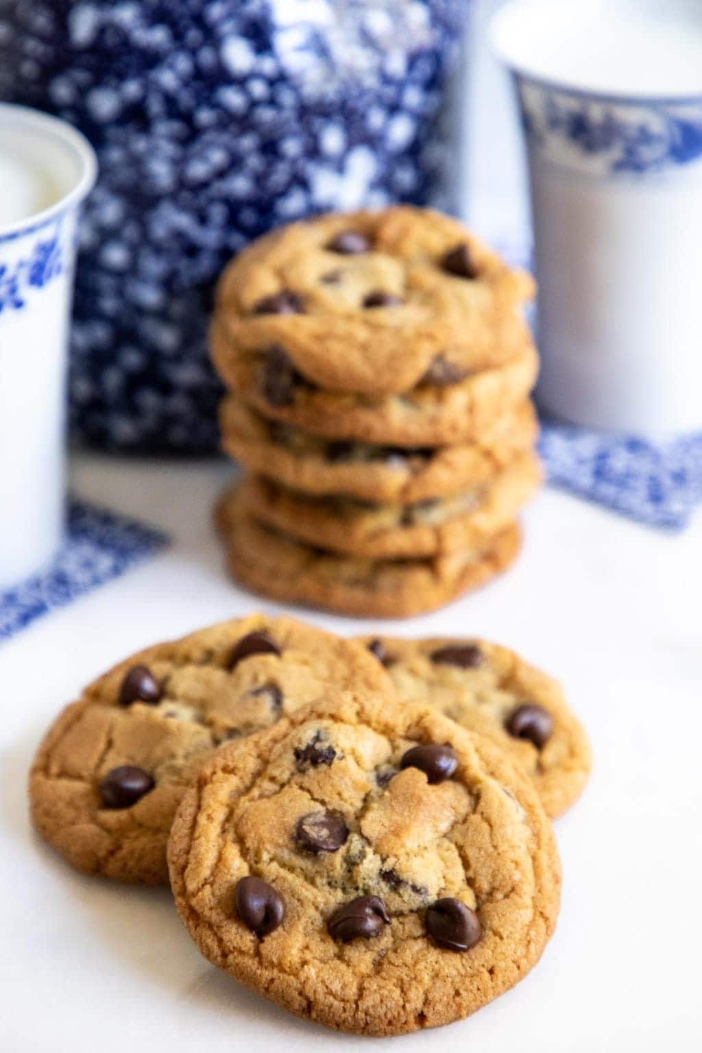Vertical photo of a batch of The BEST Chocolate Chip Cookies with a stack of them in the background with glasses of milk.