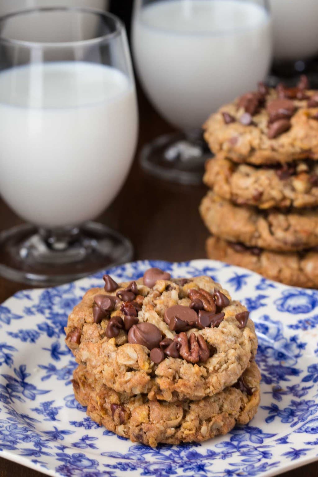 Vertical picture of Toffee Cowboy Cookies on a blue and white plate with glasses of milk.