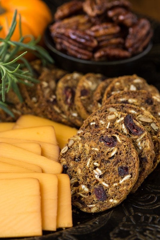 Closeup photo of an appetizer platter of Copycat Trader Joe's Pumpkin Cranberry Crisps with cheese, mini-pumpkins and pecans, garnished with a sprig of rosemary.