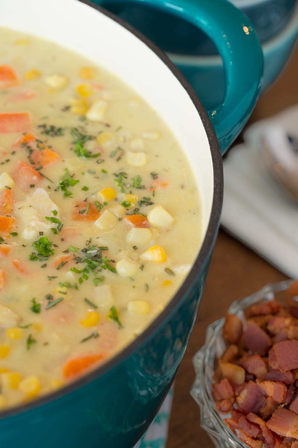 Vertical close up picture of turkey corn chowder in a turquoise dutch oven with a small dish of bacon 