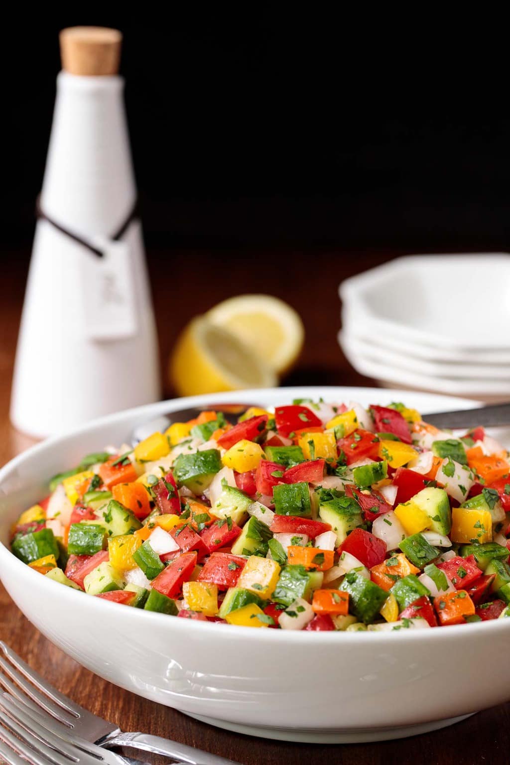 Photo of a white bowl filled with Turkish Tomato Cucumber Salad. A bottle of dressing, lemons and serving bowls are in the background.