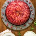 Vertical overhead picture of Upside Down Plum Cake on a platter with white dessert plates and flowers