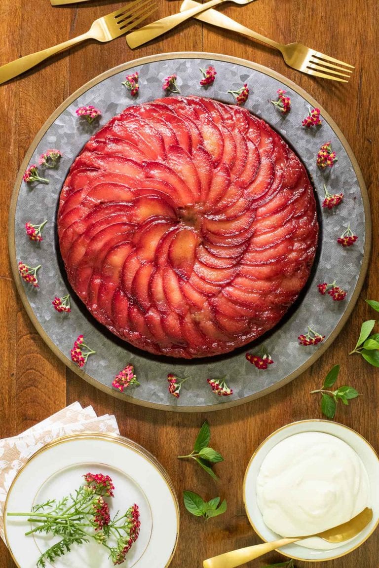Vertical overhead picture of Upside Down Plum Cake on a platter with white dessert plates and flowers