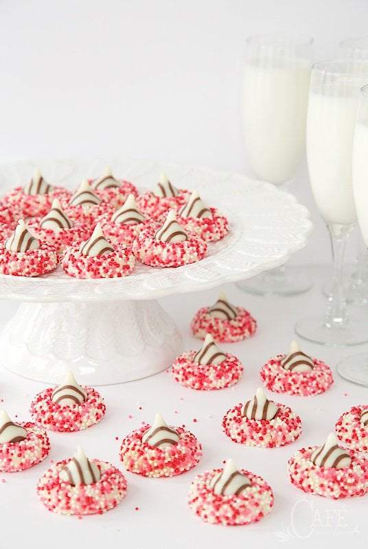 White on white photo of a batch of Easy Valentine Shortbread Cookies on a pedestal platter surrounded by additional cookies and tall glasses of milk.