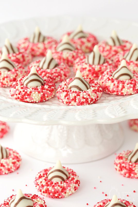 Photo of a white pedestal platter filled with Easy Valentine Shortbread Cookies. Cookies are spread on the table in the foreground of the photo too.