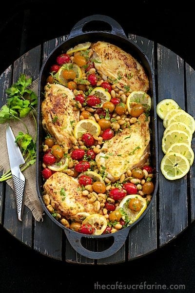 Overhead photo of a black cast iron pan filled with Mediterranean Roasted Chicken Breasts on a round wooden table with cilantro and lemon slices next to the pan.
