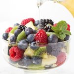 Vertical photo of a glass dish filled with fresh fruit with Limoncello Syrup being poured over the top of the berries.